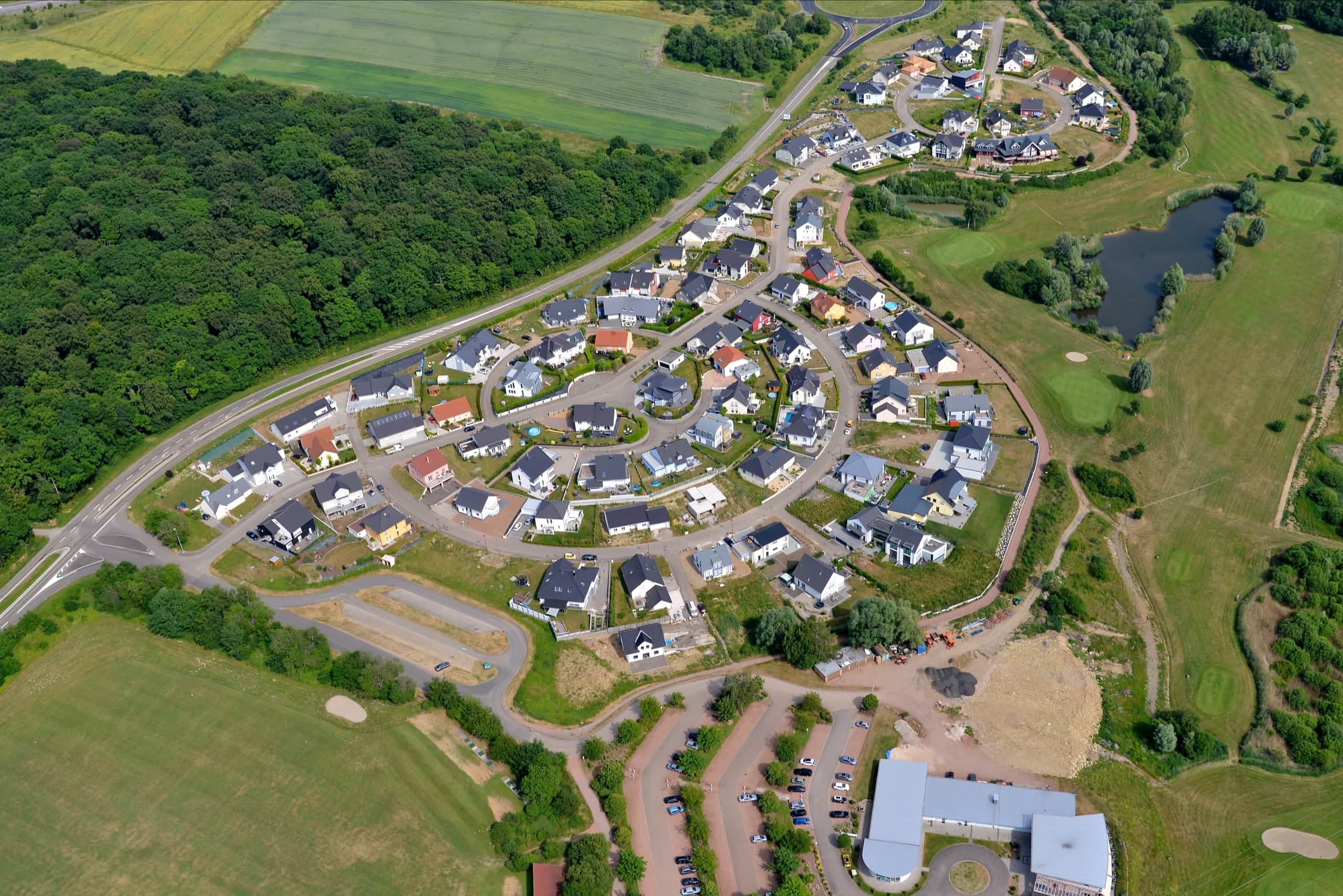 Luftbild eines erschlossenen Wohngebiets mit Ringstraße, Einfamilienhäusern und Regenrückhaltebecken am Waldrand